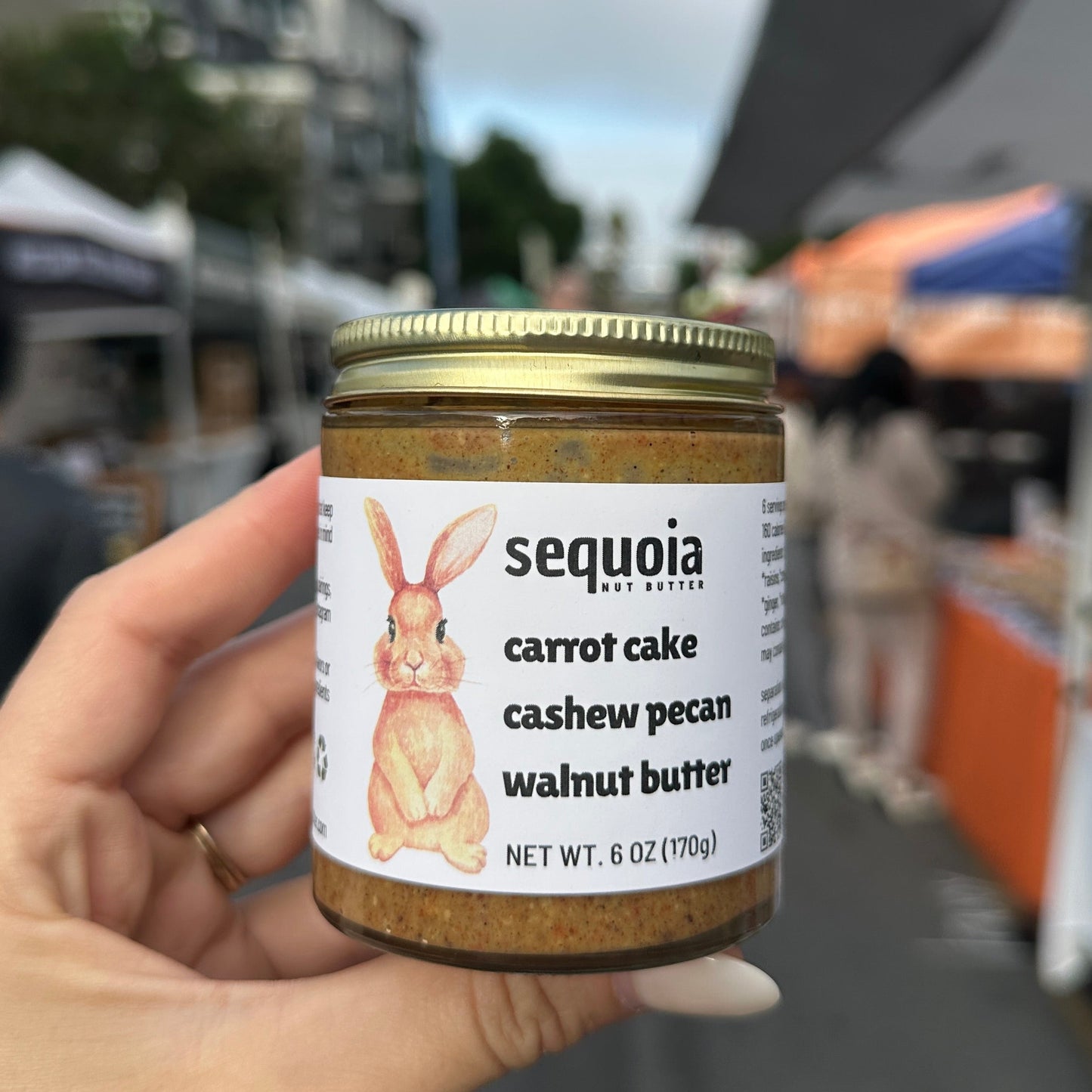 holding a jar of Sequoia Carrot Cake Cashew Pecan Walnut Butter at an outdoor event.