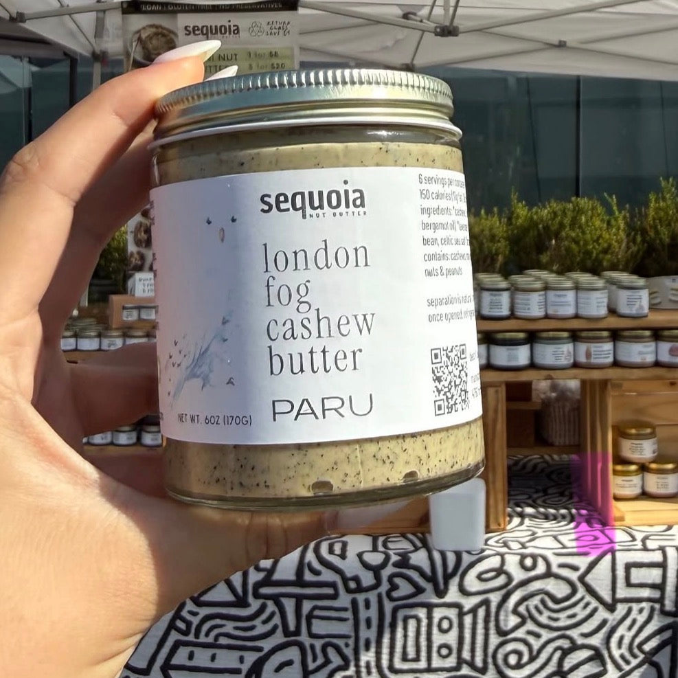 A hand holds a glass jar of Sequoia London Candle Co. London Fog Cashew Butter against an outdoor market background with other jars on display.