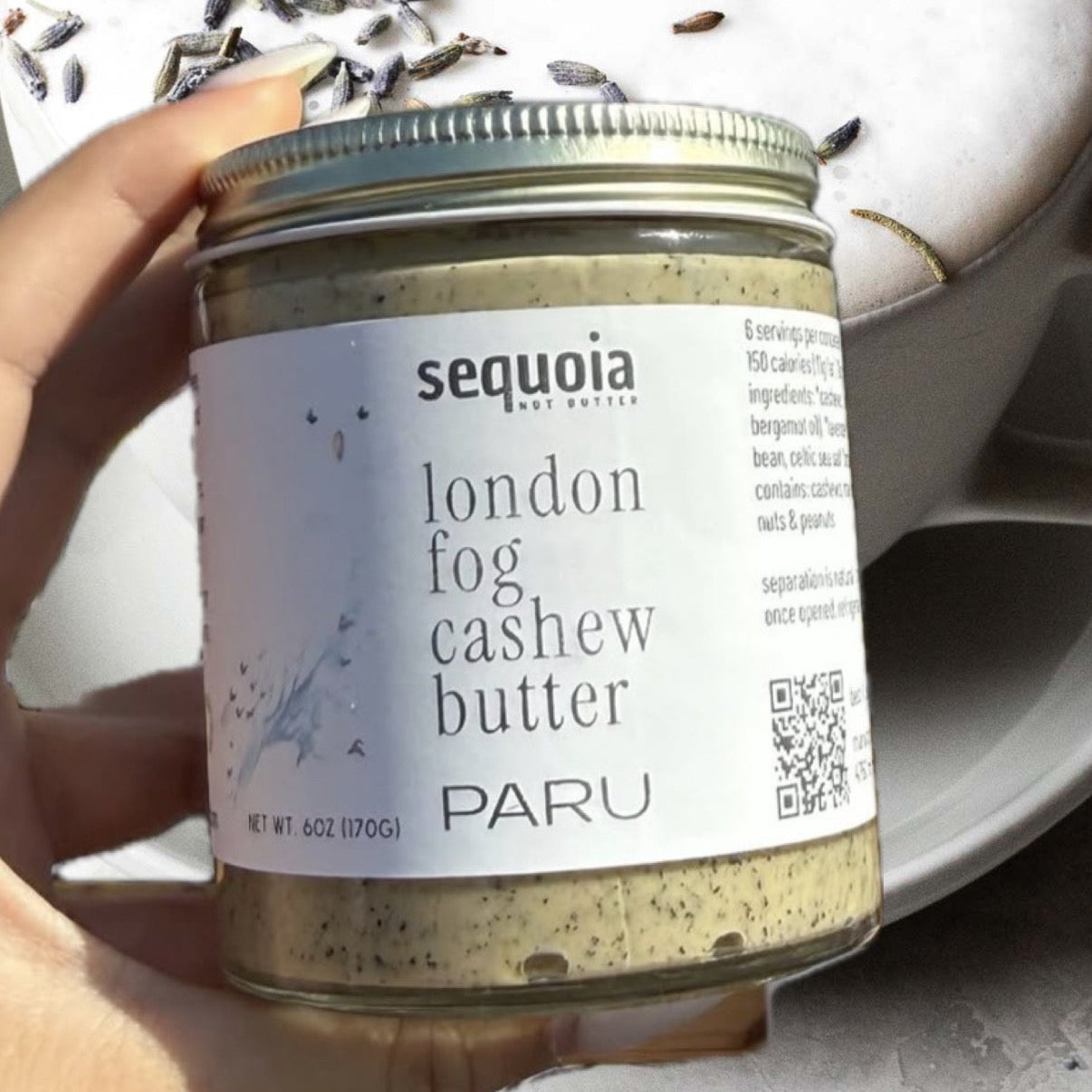 A hand holds a glass jar of Sequoia London Fog Cashew Butter against a styled background of a white ceramic bowl with dried lavender and a mug. Text at the top reads 'this weeks test batch!! thai earl grey from @parutea'.